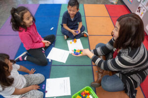 Teacher sitting with young students on rainbow rug.