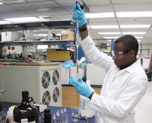 A black man wearing a lab coat and safety goggles uses chemistry equipment in a lab