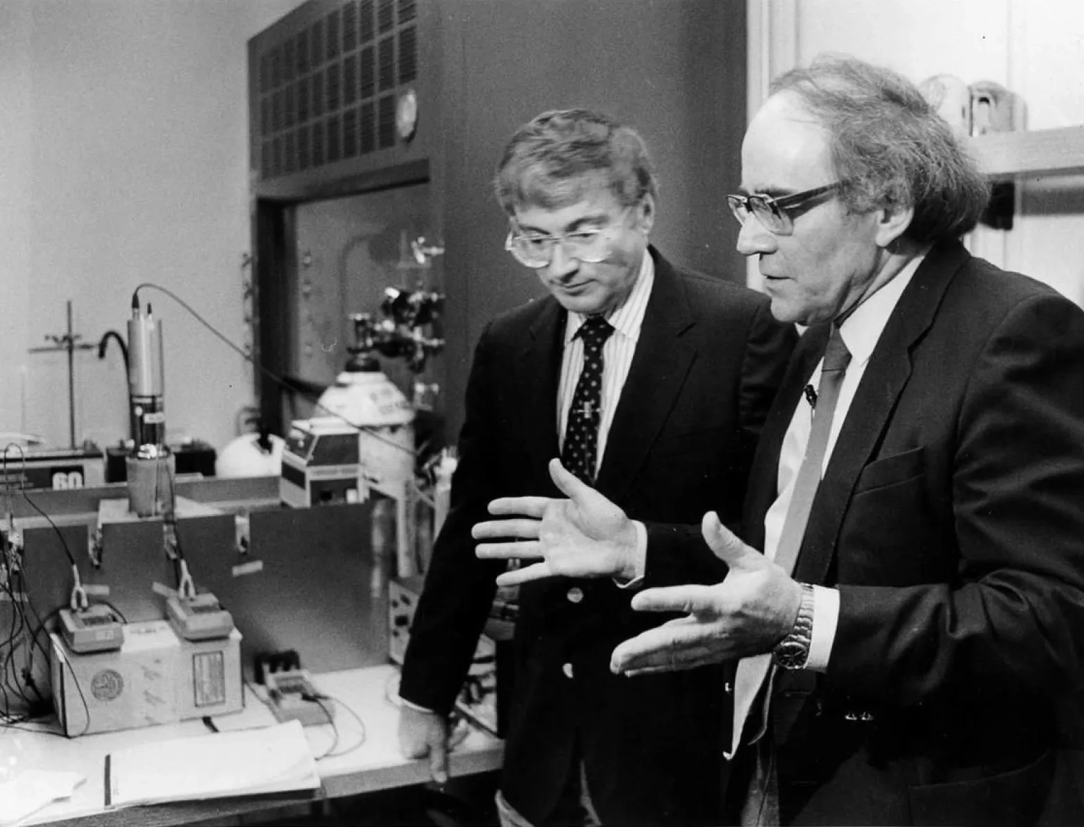 Two white men in suits talk near old fashioned chemistry equipment