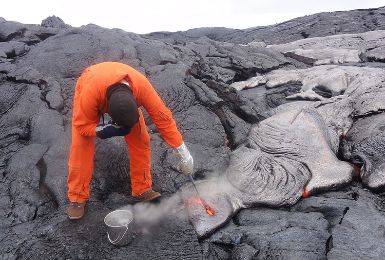 a man wearing protective clothing is reaching over lava flows to collect a sample