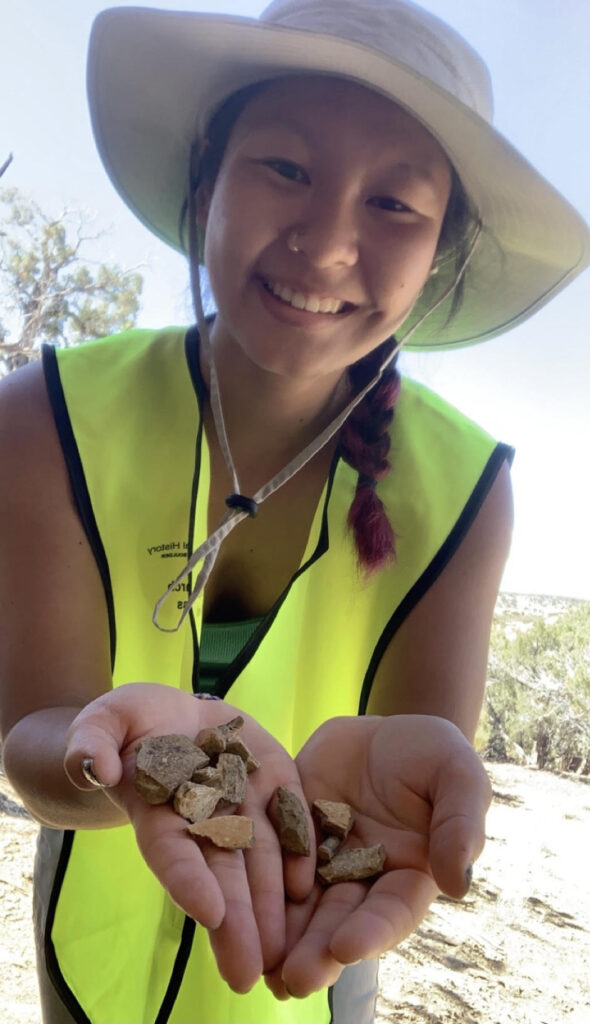 PhD student Kayli Stowe looks at the camera while holding out her hands which show several small fossils. She is wearing a hat and work vest.